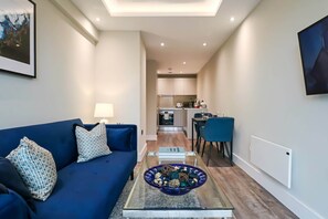 Un salon moderne avec un canapé bleu, une table basse en verre agrémentée d’objets décoratifs, un coin repas avec une chaise bleue, et une cuisine en arrière-plan.