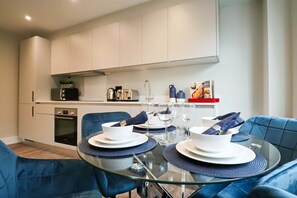 Une cuisine moderne avec des armoires blanches, un four encastré et un coin repas doté de chaises à revêtement bleu et d’une table en verre, le tout disposé pour deux personnes.