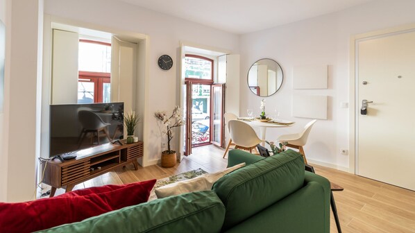 A modern living room with a flat-screen TV, green sofas, a dining area with a table and chairs, and a large mirror on the wall.