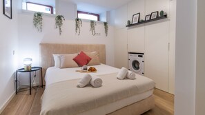 Un dormitorio con cama, almohadas, una mesita de noche con lámpara, una pequeña mesa con comida, una lavadora y un estante con fotografías.