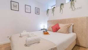 A bedroom with a bed, towels, a tray with croissants, framed pictures on the wall, and hanging plants.