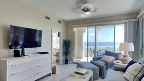 A modern living room with a flat-screen TV, a white dresser, a bench, a sofa, and a view of the ocean through large sliding doors.