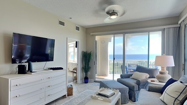 A modern living room with a flat-screen TV, a white dresser, a bench, a sofa, and a view of the ocean through large sliding doors. - Surf Club by VTrips (Palm Coast)