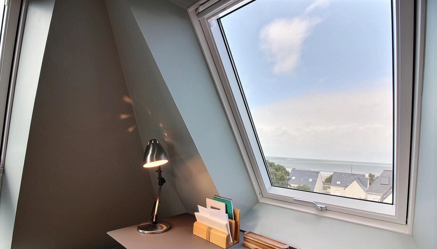 Una scrivania angolare con lampada da tavolo, una finestra che si affaccia su edifici e mare, e un portapenne.