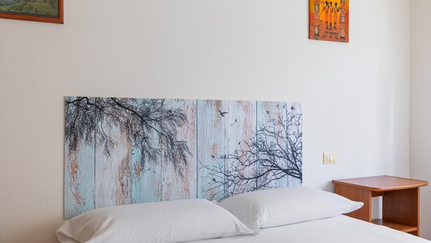 A bedroom with a bed, white pillows, a wooden nightstand, and a wall with a large painting of trees. - Urban Cozy Apartment by Wonderful Italy (Bologna)
