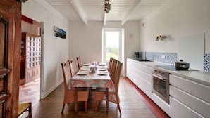 Un coin repas avec une table dressée pour six personnes, entourée de chaises en osier, une cuisine avec des meubles blancs et un dosseret carrelé, et un plafond en bois avec des poutres apparentes.