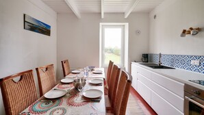 Un coin repas avec une table dressée pour quatre personnes, entourée de chaises en osier, d’une nappe à motifs et offrant une vue sur l’extérieur par une fenêtre.