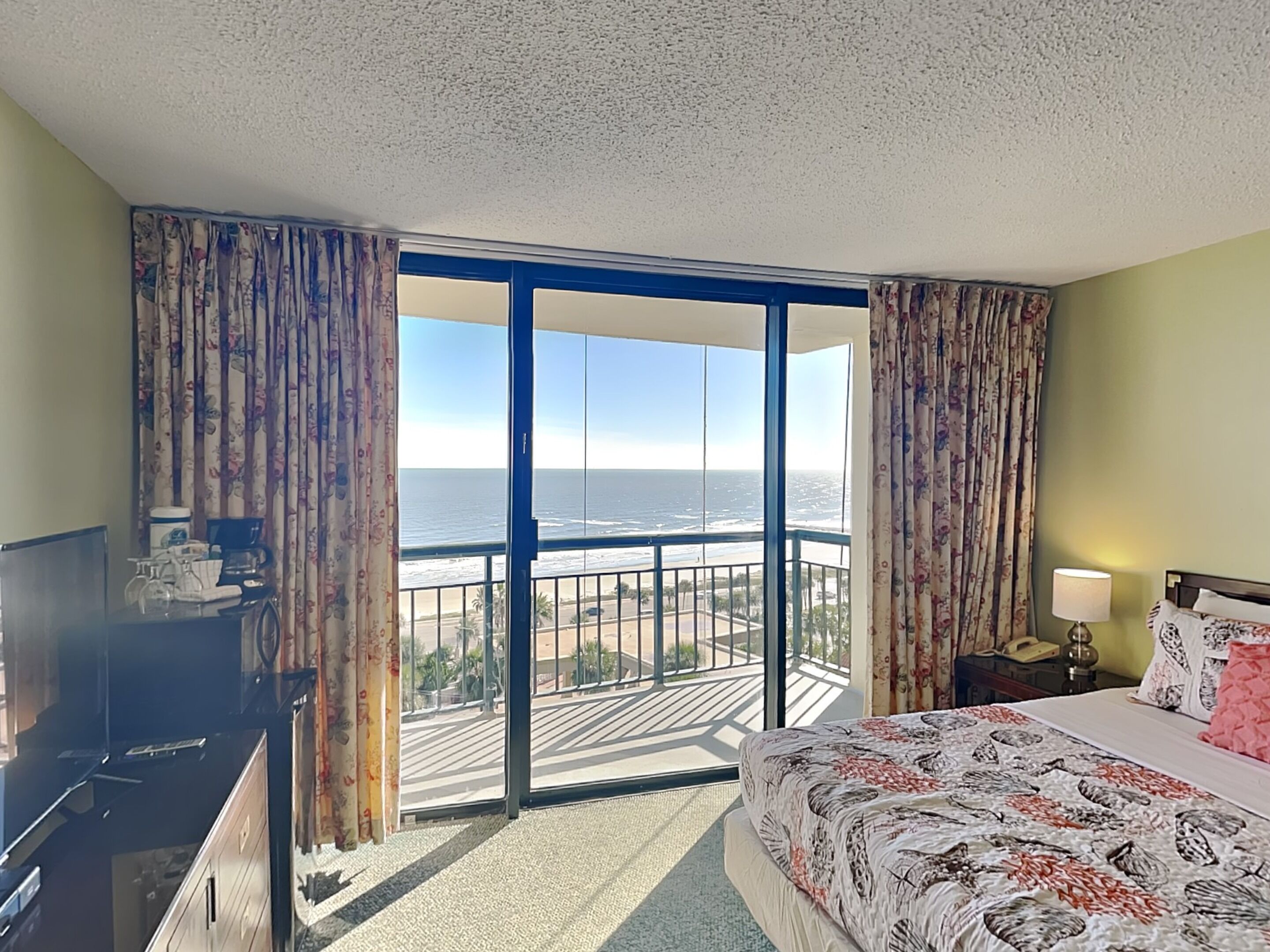 Una habitación de hotel con un balcón amplio, una cama con edredón estampado con motivos florales, un escritorio con computadora y una mesita de noche con lámpara.