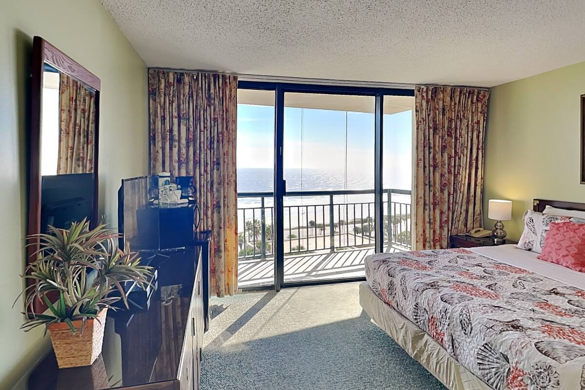 A hotel room with a bed, a desk with a TV, a potted plant, and a balcony with a view of the sea.