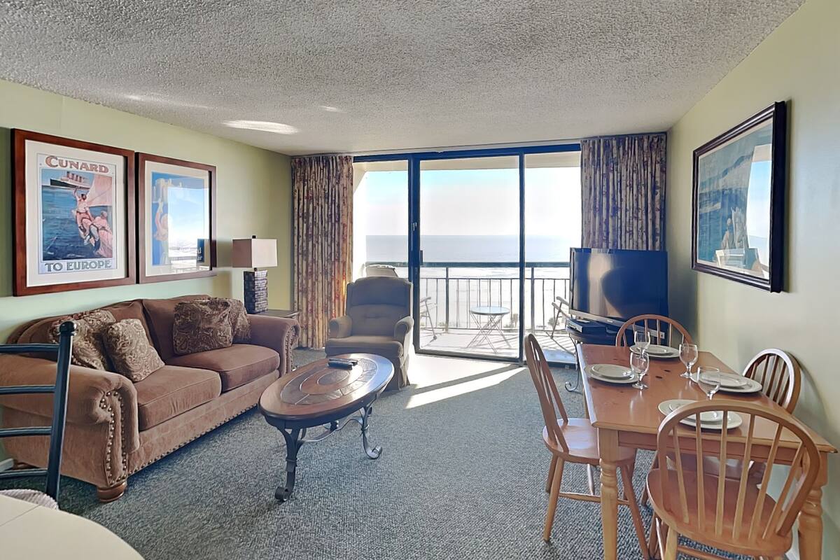 A hotel room with a balcony, a sofa, a coffee table, a TV, and a dining table set for two.