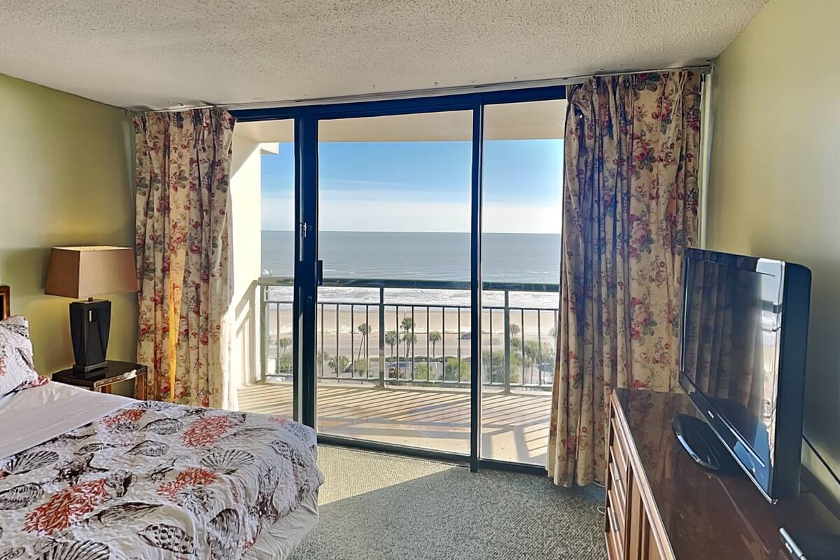A hotel room with a bed, a television, a desk, and a view of the beach and ocean.