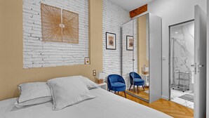 Un dormitorio moderno con pared de ladrillo blanco, una cama con ropa de cama blanca, dos sillas azules y una cabina de ducha de vidrio.