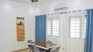 A room with a wooden table and six chairs, a painting on the wall, and a window with blue curtains. - L'Oasis Mid-Century (Cotonou)