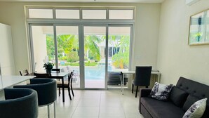 Ein modernes Wohnzimmer mit Sofa, Sesseln, einem Esstisch und Blick auf Pool und Grünflächen durch große Glasfenster.