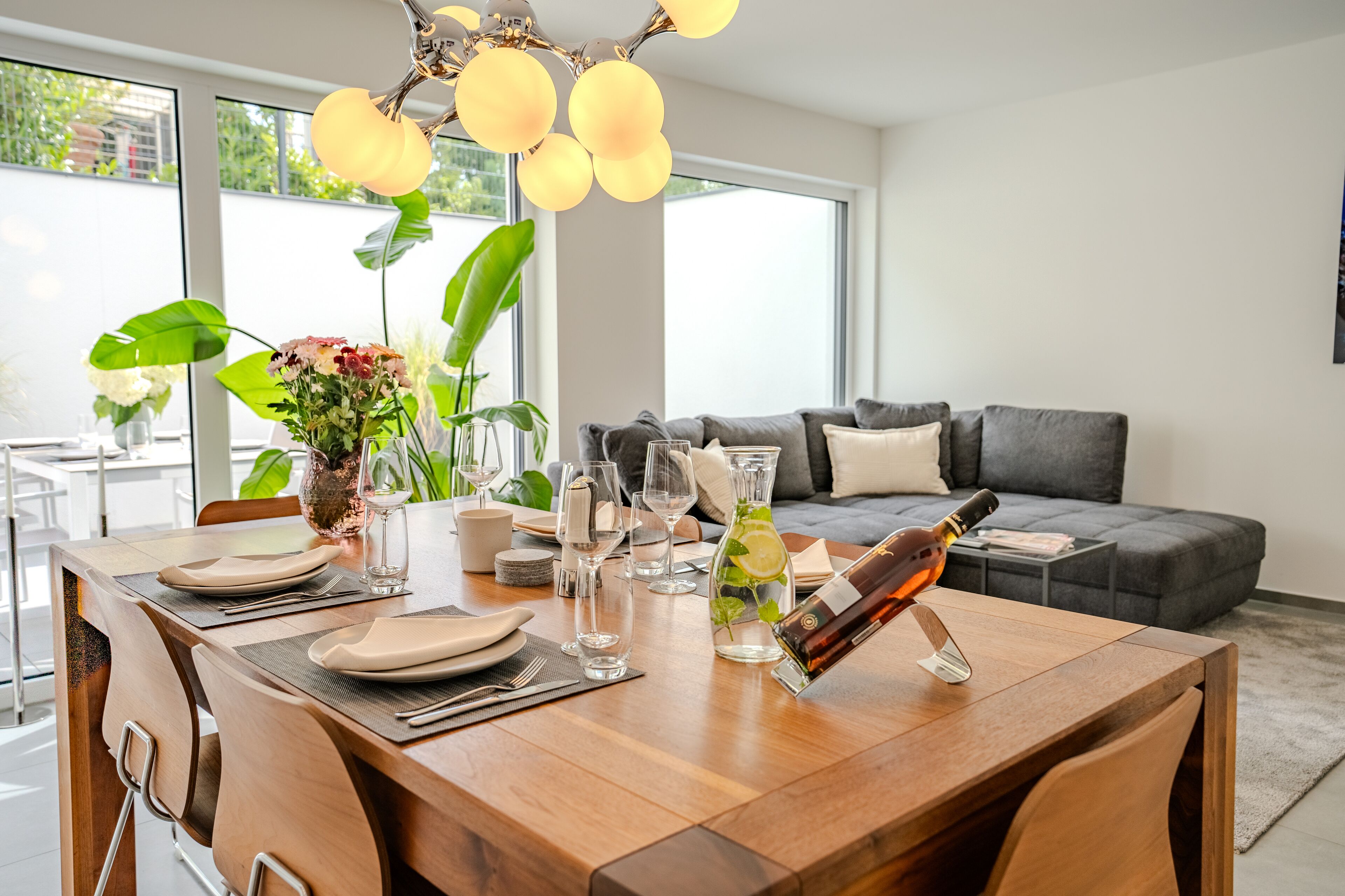 Une table à manger dressée avec des assiettes, des verres et une bouteille de champagne, surmontée d’un lustre moderne.