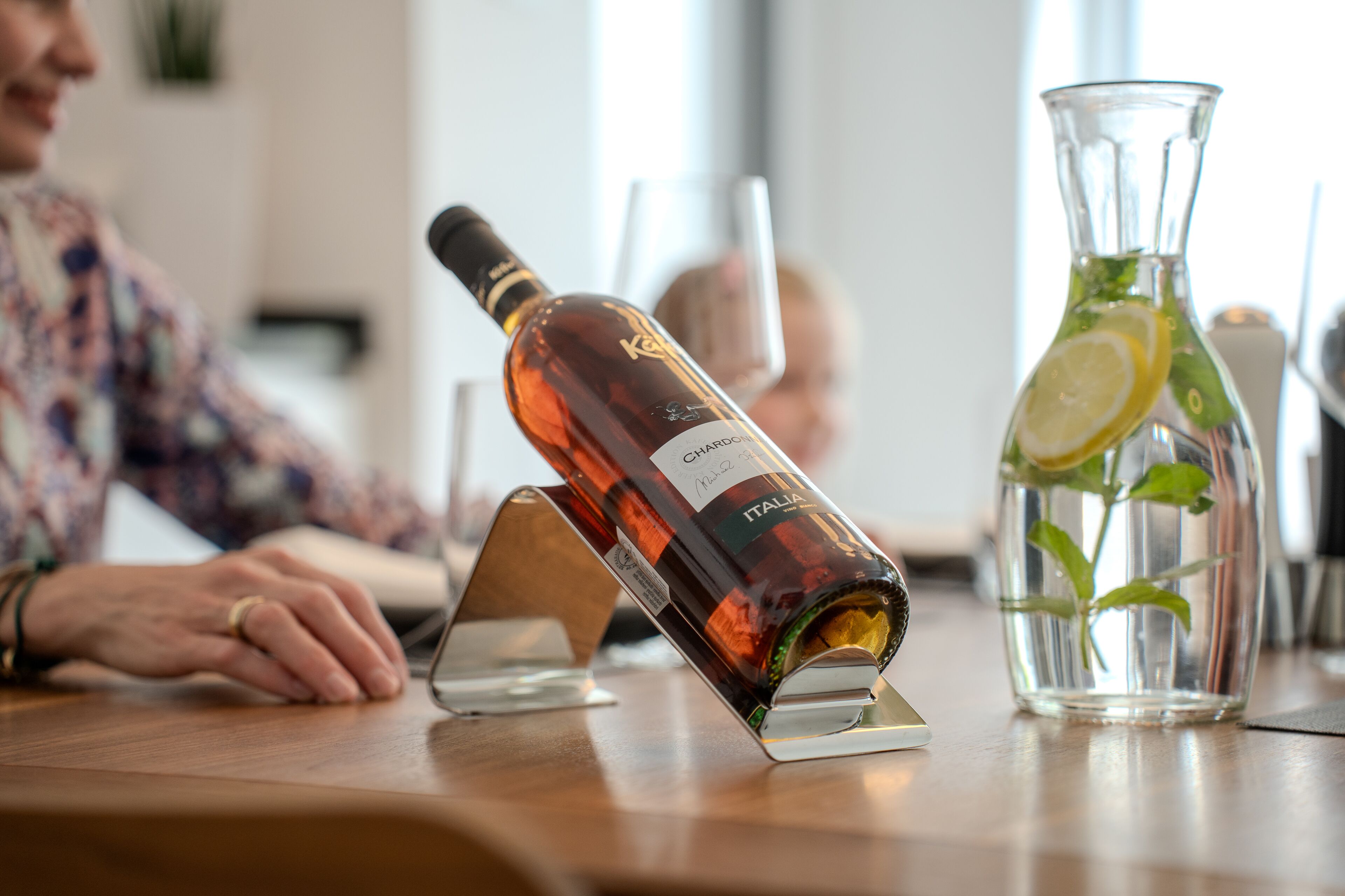En flaska Chianti Classico-vin står på ett stativ, bredvid en glasbringa med en citronskiva och myntablad, och i bakgrunden syns en person.