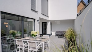 A modern outdoor patio with a dining table set for four, surrounded by white chairs. - NEU Luxuswohnung in Waldrandlage Nähe Köln Messe! (Leverkusen)