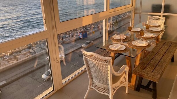 A balcony with a table set for dining, overlooking a coastal area with a view of the sea and a distant cityscape.
