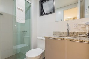 A modern bathroom with a glass shower enclosure, a white toilet, a granite countertop sink, and a mirror above the sink. - 1 room apartment versatile (Curitiba)