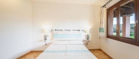 Quarto com cama, duas luminárias de cabeceira, janela com cortinas e vista para o verde lá fora.