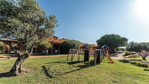 A grassy playground with a slide and swings, surrounded by trees and buildings. - Residence Baia dei Mirti (Aglientu)
