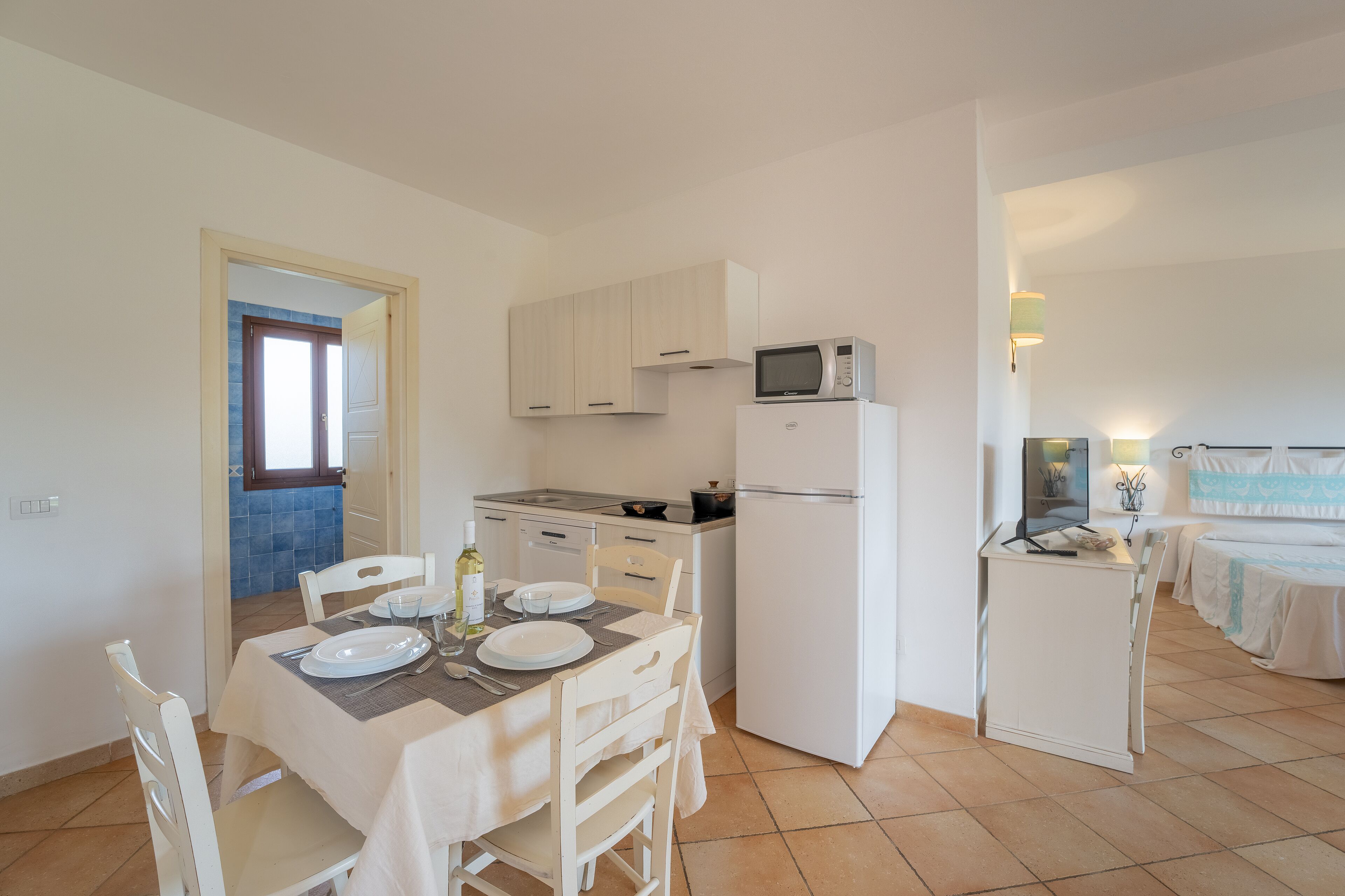 A compact kitchen with white cabinets, a microwave, and a refrigerator. There is a dining table set with plates, cutlery, and a bottle of wine.
