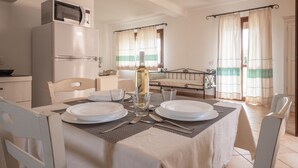 A dining area with a table set for two, a microwave above the refrigerator, and a bottle of wine on the table. - Residence Baia dei Mirti (Aglientu)