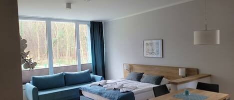 Un dormitorio moderno con cama, sofá, mesa de comedor y un ventanal con vistas a árboles.