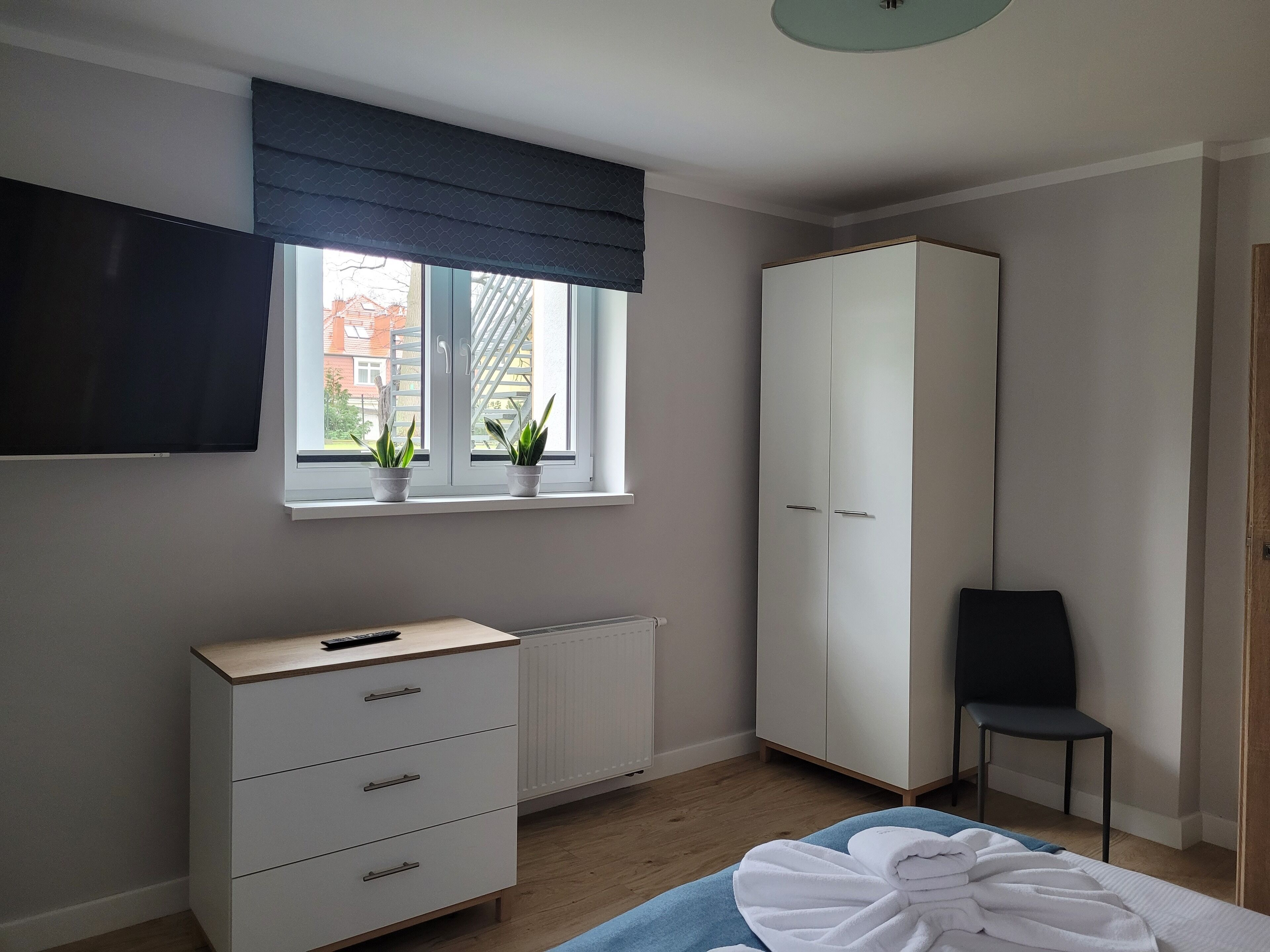 Un dormitorio con cama, armario, escritorio con silla, radiador y ventana con plantas.