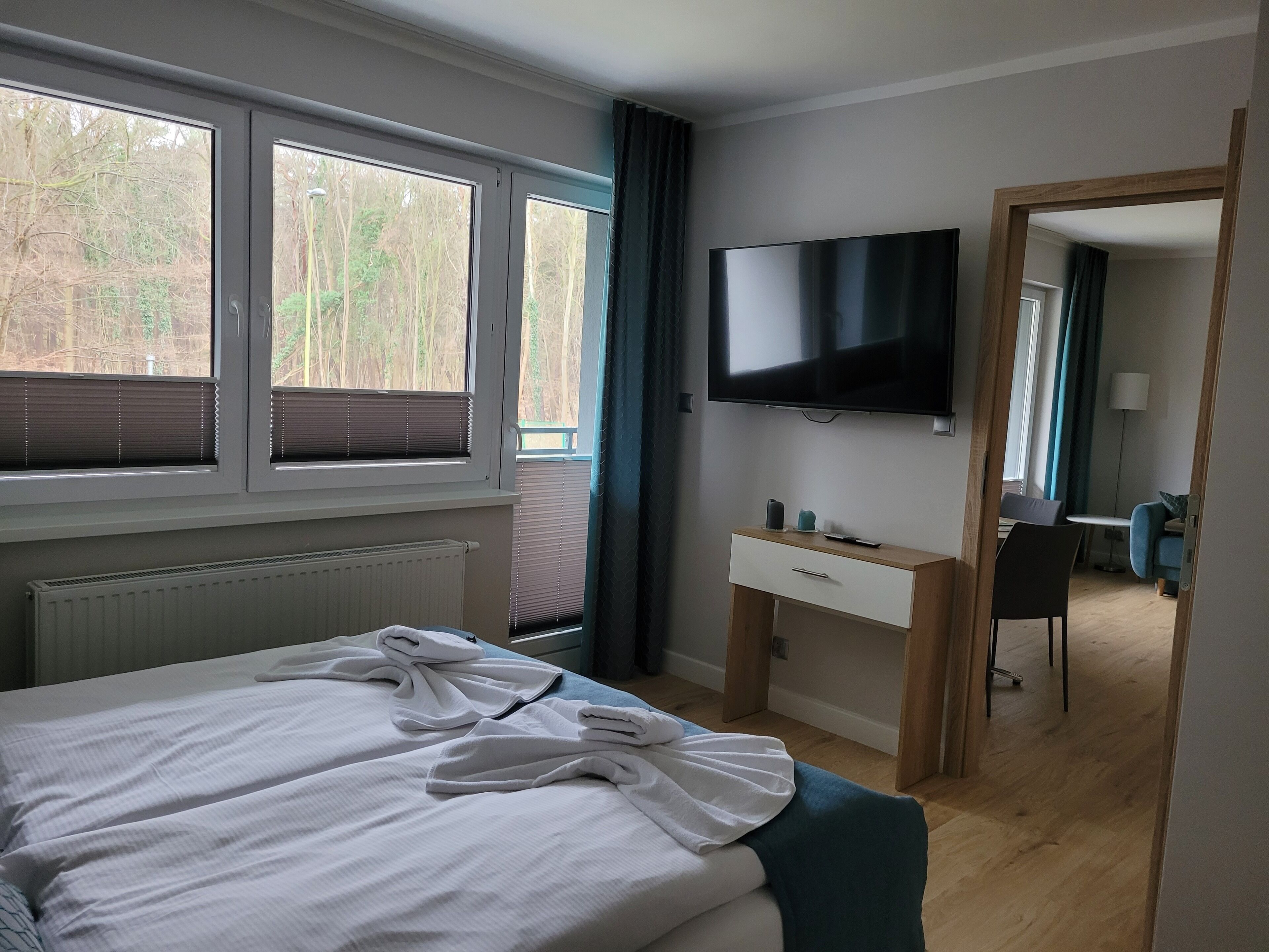 Habitación de hotel con cama, escritorio, silla, televisión y vistas a árboles desde las ventanas.
