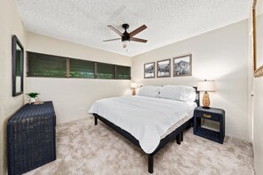 Une chambre à coucher avec un lit, un ventilateur de plafond, une fenêtre avec des stores, deux lampes de chevet et des tableaux encadrés accrochés au mur.