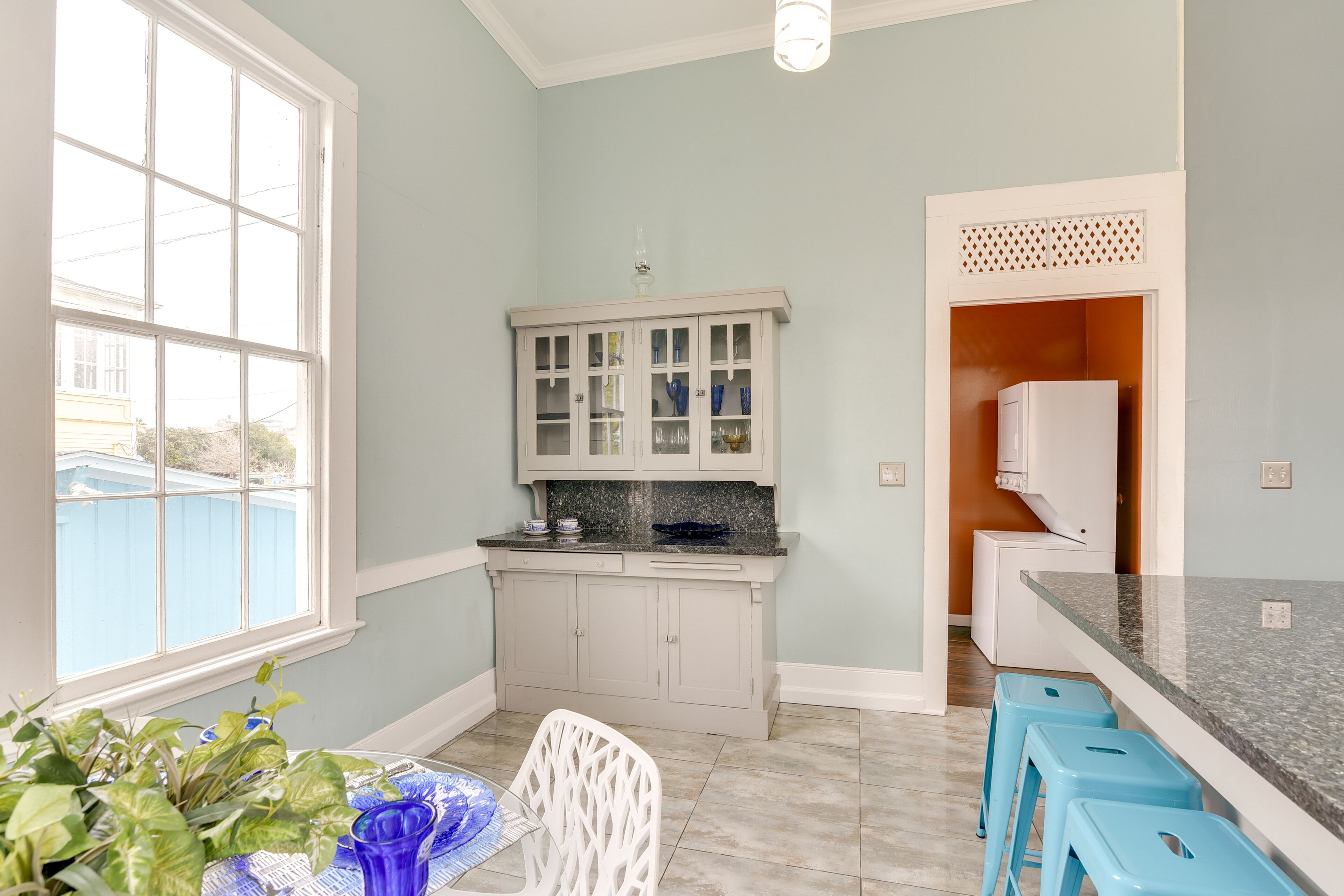 A kitchen with a window, a white cabinet with glass doors, a countertop, and blue stools.