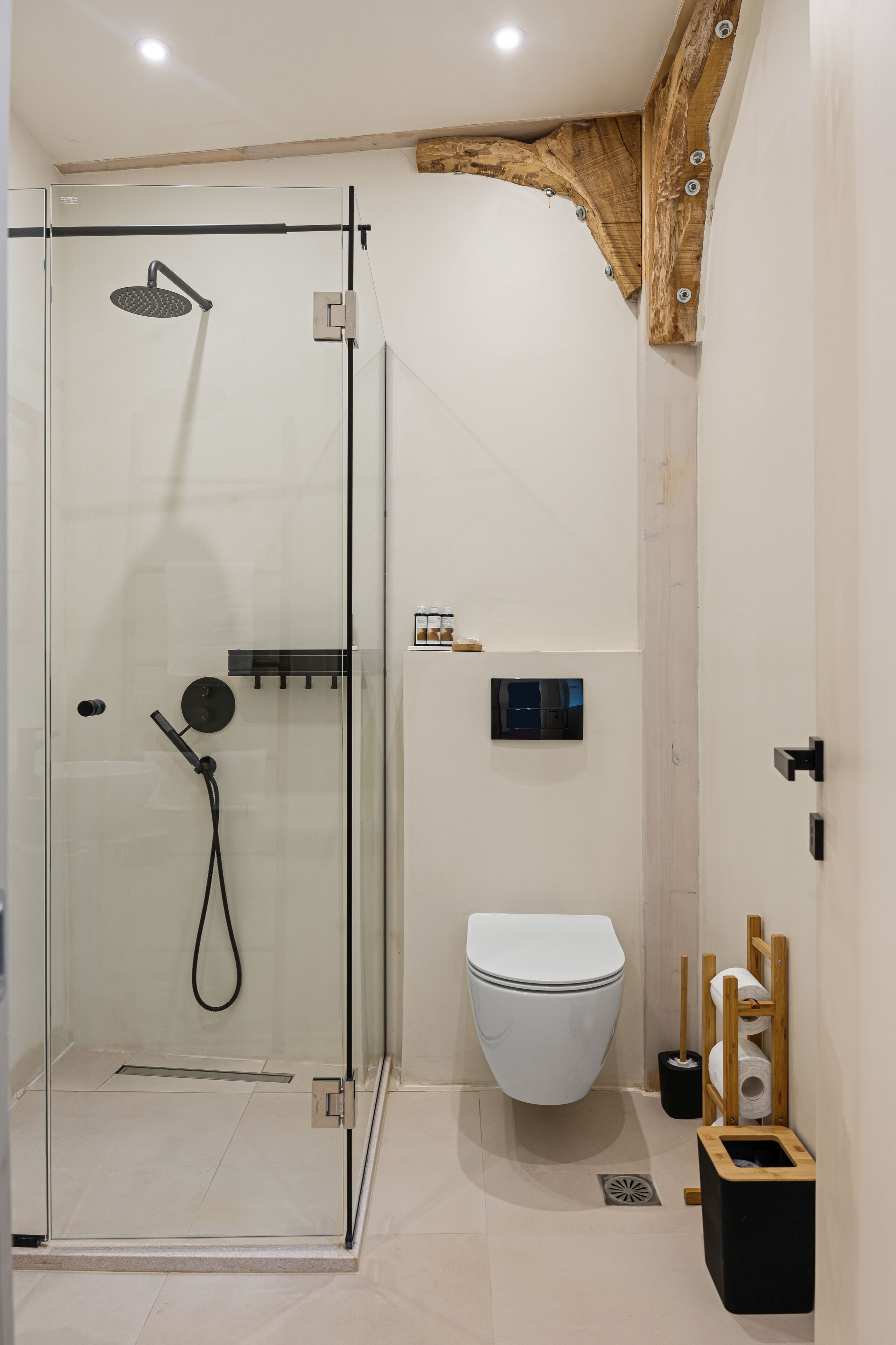 A modern bathroom with a glass-enclosed shower, a white toilet, and a wooden shelf with toiletries.