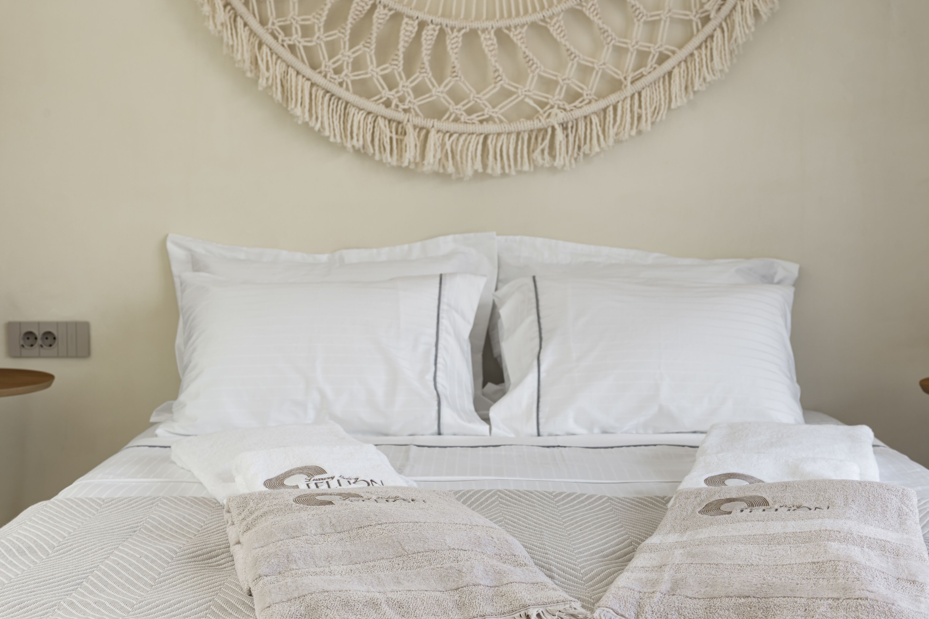 Una cama bien hecha con sábanas blancas y dos almohadas, un colgador de macramé en la pared, y una mesita de noche con una lámpara.