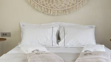 Una cama bien hecha con sábanas blancas y dos almohadas, un colgador de macramé en la pared, y una mesita de noche con una lámpara.