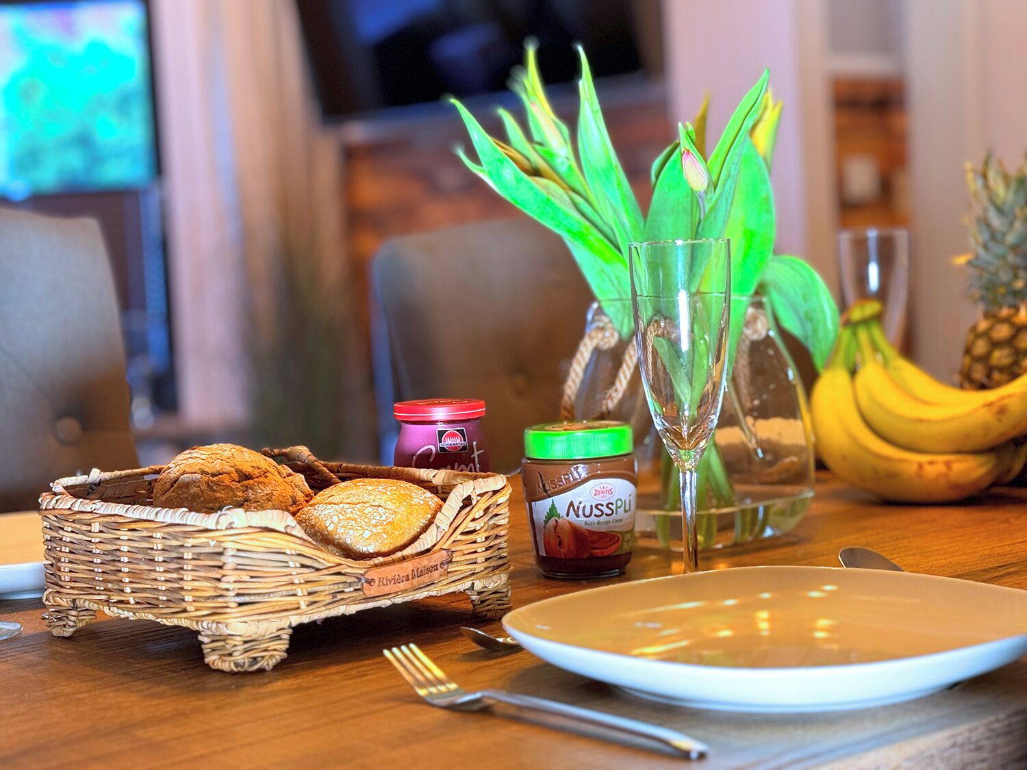 Une table à manger avec un panier en osier contenant du pain, un pot de Nutella, un verre d’eau, une banane et un ananas.