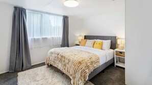 Ein modernes Schlafzimmer mit einem großen Bett, grauen Vorhängen, einem weißen Teppich und einem Nachttisch mit einer Lampe.