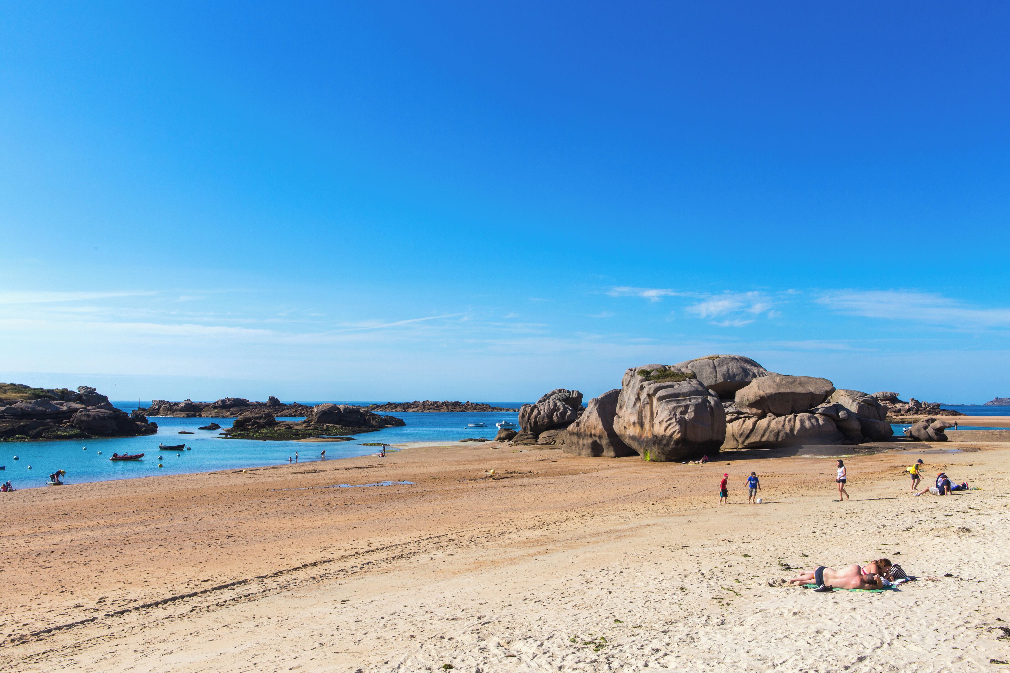 Ein Strand mit großen Felsen, klarem, blauem Wasser und sonnenbadenden Personen.