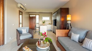 Een moderne woonkamer met een bank, fauteuils, een salontafel met een schaal gebakjes, een potplant en een keuken op de achtergrond.
