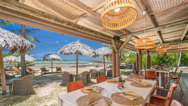 Een terras met rieten meubels, rieten parasols en uitzicht op het strand.