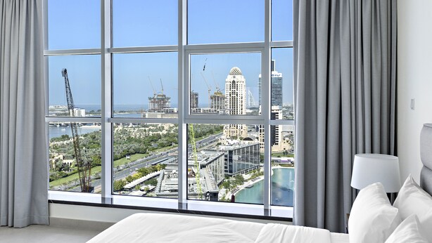 A hotel room with a large window offering a city view, a bed, and a nightstand with a lamp. - WelHome - Breathtaking Partial Sea View 2BR Close to Marina  (Dubai)