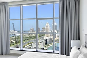 A hotel room with a large window offering a city view, a bed, and a nightstand with a lamp. - WelHome - Breathtaking Partial Sea View 2BR Close to Marina  (Dubai)