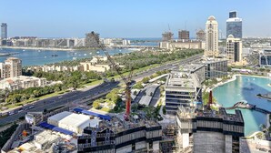 A cityscape with a prominent waterfront, including a marina with boats, modern high-rise buildings, and a large construction site with cranes. - WelHome - Breathtaking Partial Sea View 2BR Close to Marina  (Dubai)