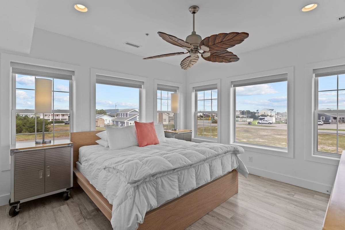 A bedroom with a bed, a ceiling fan, large windows, and a view of the outside.
