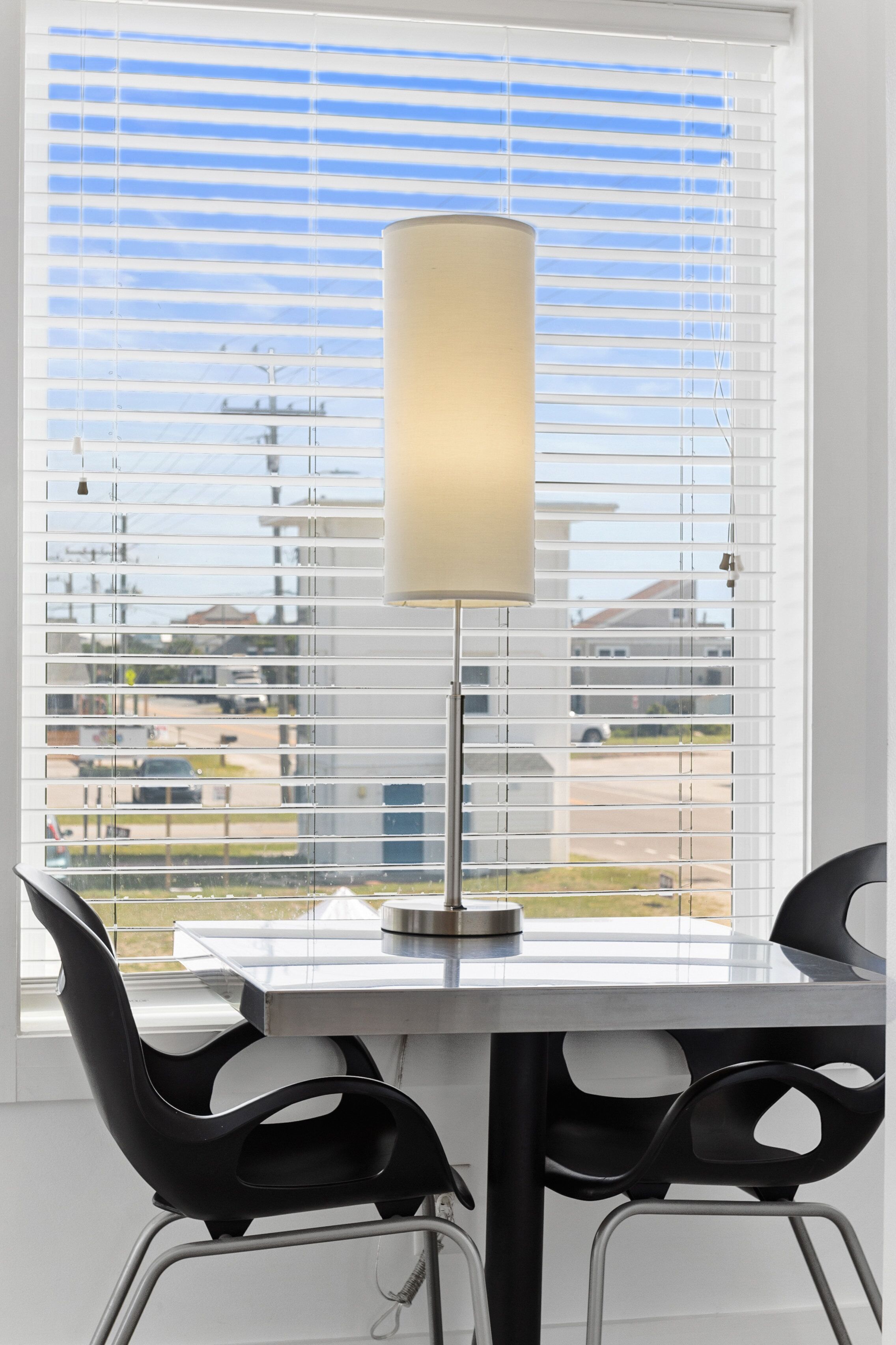 A modern dining area with a table and two chairs, a lamp, and a view through window blinds.