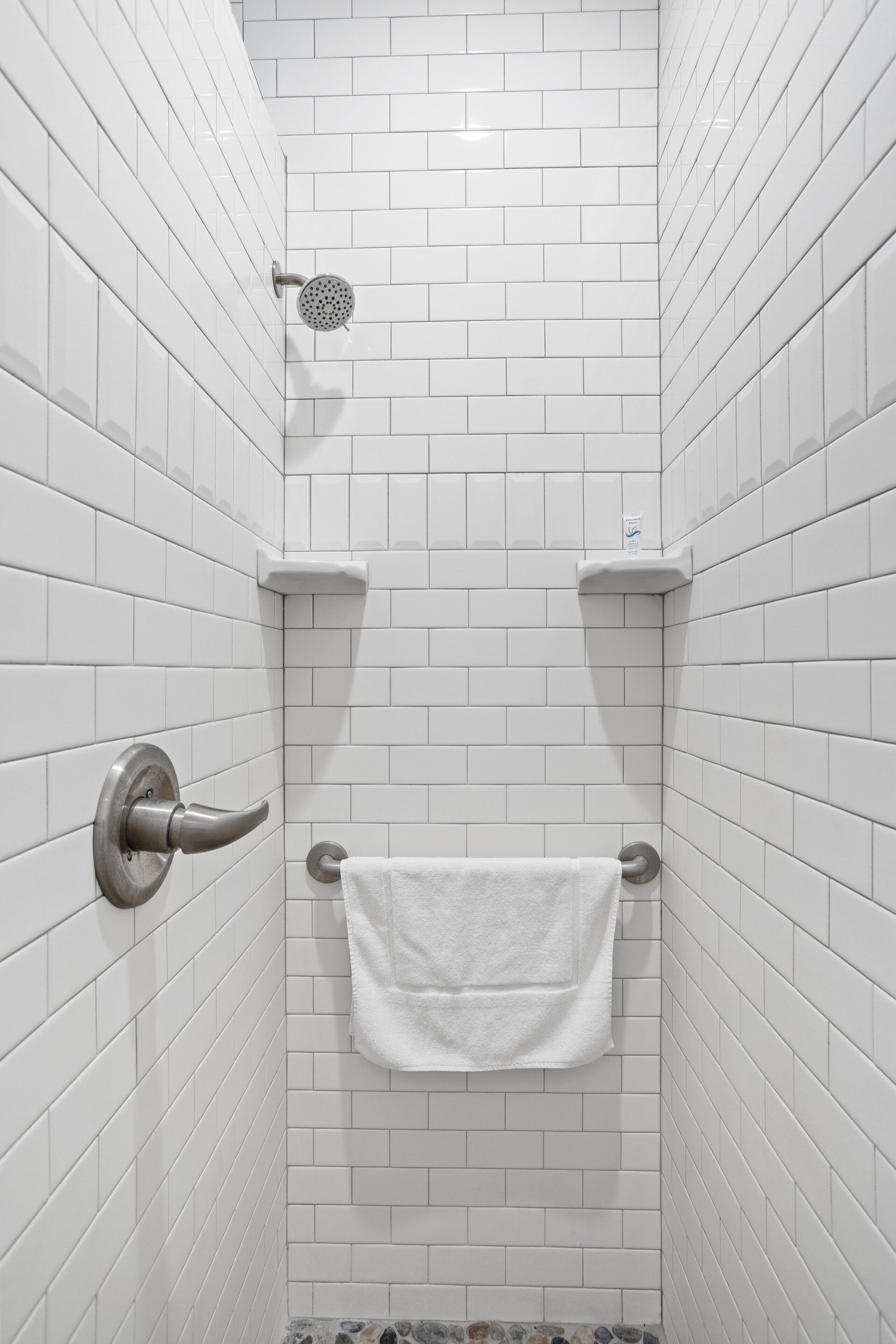 A small, white-tiled shower stall with a handheld showerhead, a towel rack, and a small shelf.