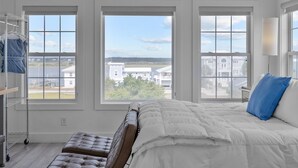A bedroom with a large bed, a bench, a metal shelf, and three windows offering a view of buildings and greenery.