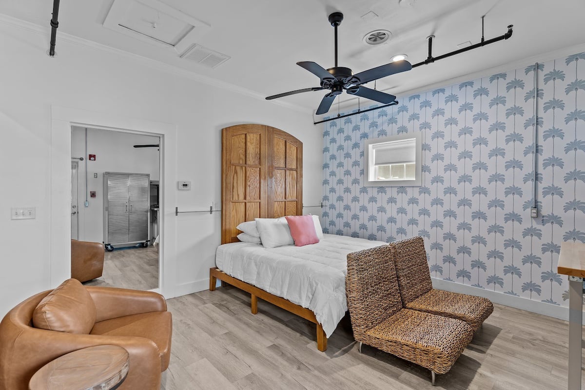 A bedroom with a bed, a wooden headboard, a ceiling fan, a patterned wall, and a door leading to a bathroom.