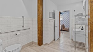 A modern bathroom with white tiled walls, a wooden column, a grab bar, a toilet, and a shower area.