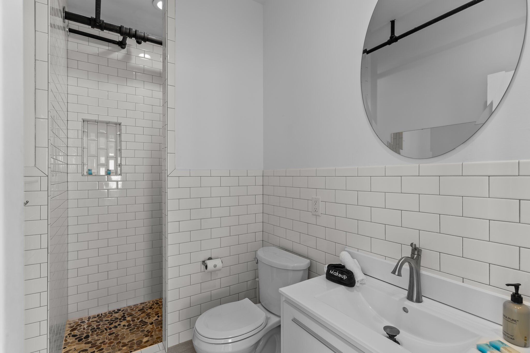 Une salle de bain moderne avec des carreaux blancs, une douche, des toilettes, un lavabo avec un miroir et une trousse de maquillage posée sur le comptoir.